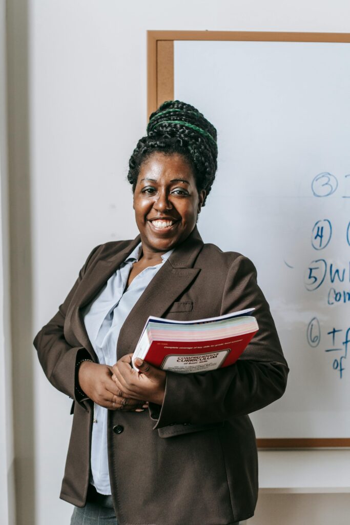 Cheerful African American female teacher wearing formal clothes standing with stacked notebooks in hands near whiteboard in classroom and looking at camera with toothy smile